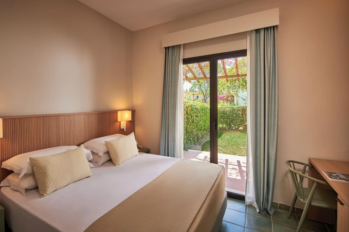 A hotel room with a bed, a desk, a chair, a lamp, and a view of a garden through a sliding door.