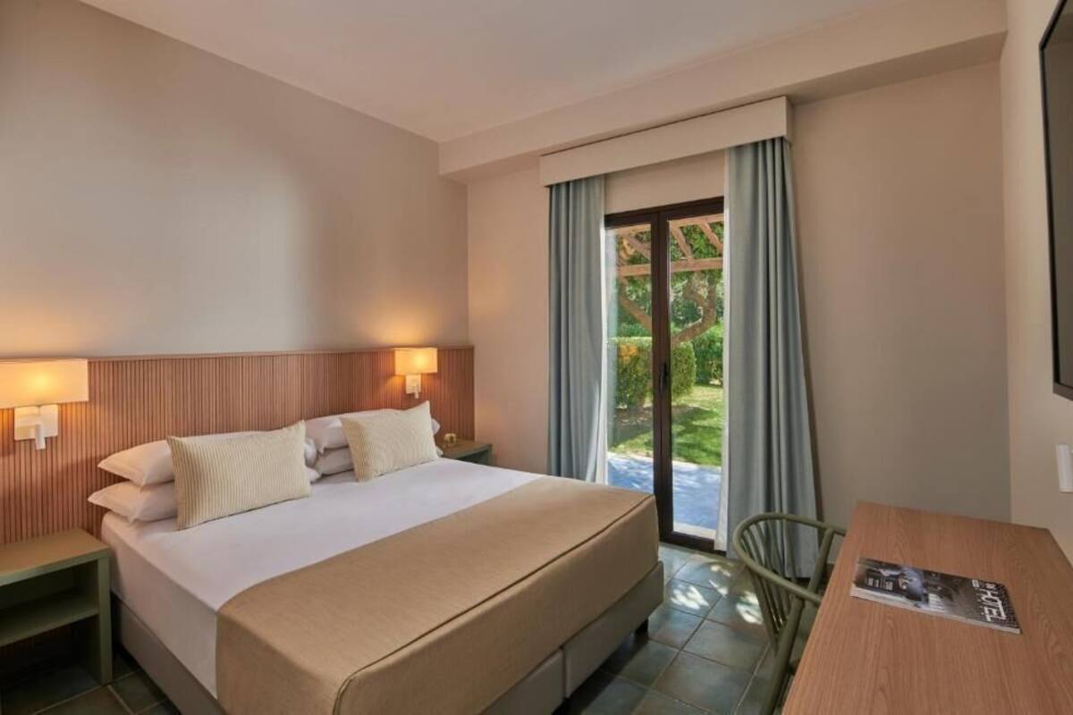 A hotel room with a bed, bedside table, lamp, window with curtains, and a view of greenery outside.