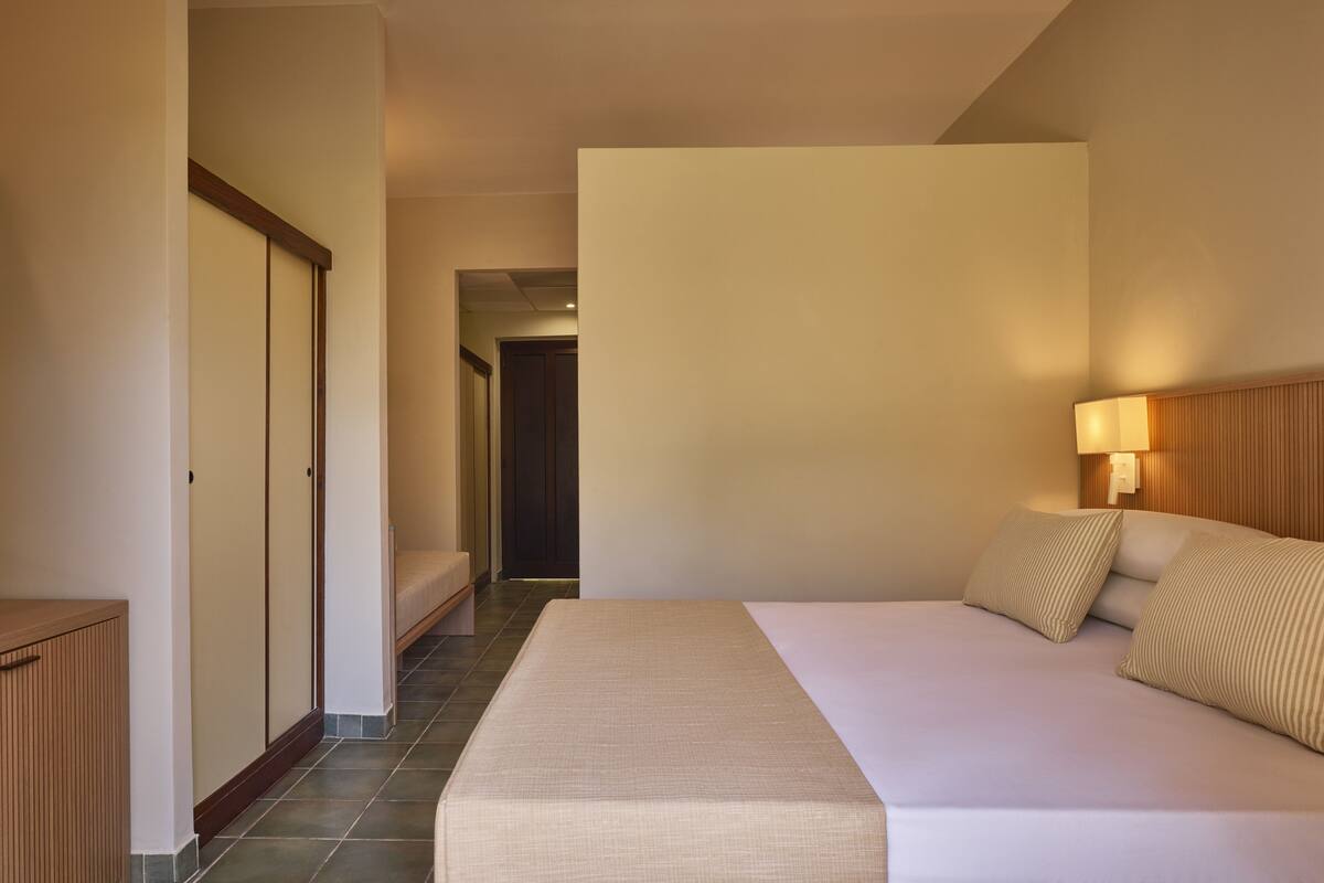 A hotel room with a bed, a bedside table, a lamp, and a view of a hallway.