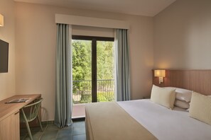 A hotel room with a bed, a desk, and a view of greenery through the window.