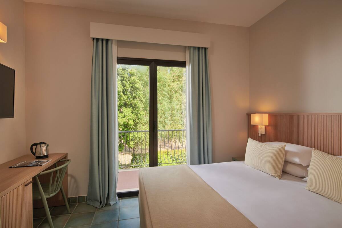 A hotel room with a bed, a desk, a chair, a window with curtains, and a view of greenery outside.