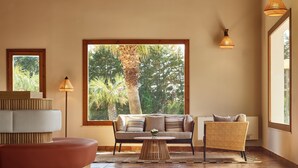 A modern living room with a sofa, a wicker chair, and a coffee table. There are large windows providing a view of trees and a palm tree. - Serenè Resort (Cutro)