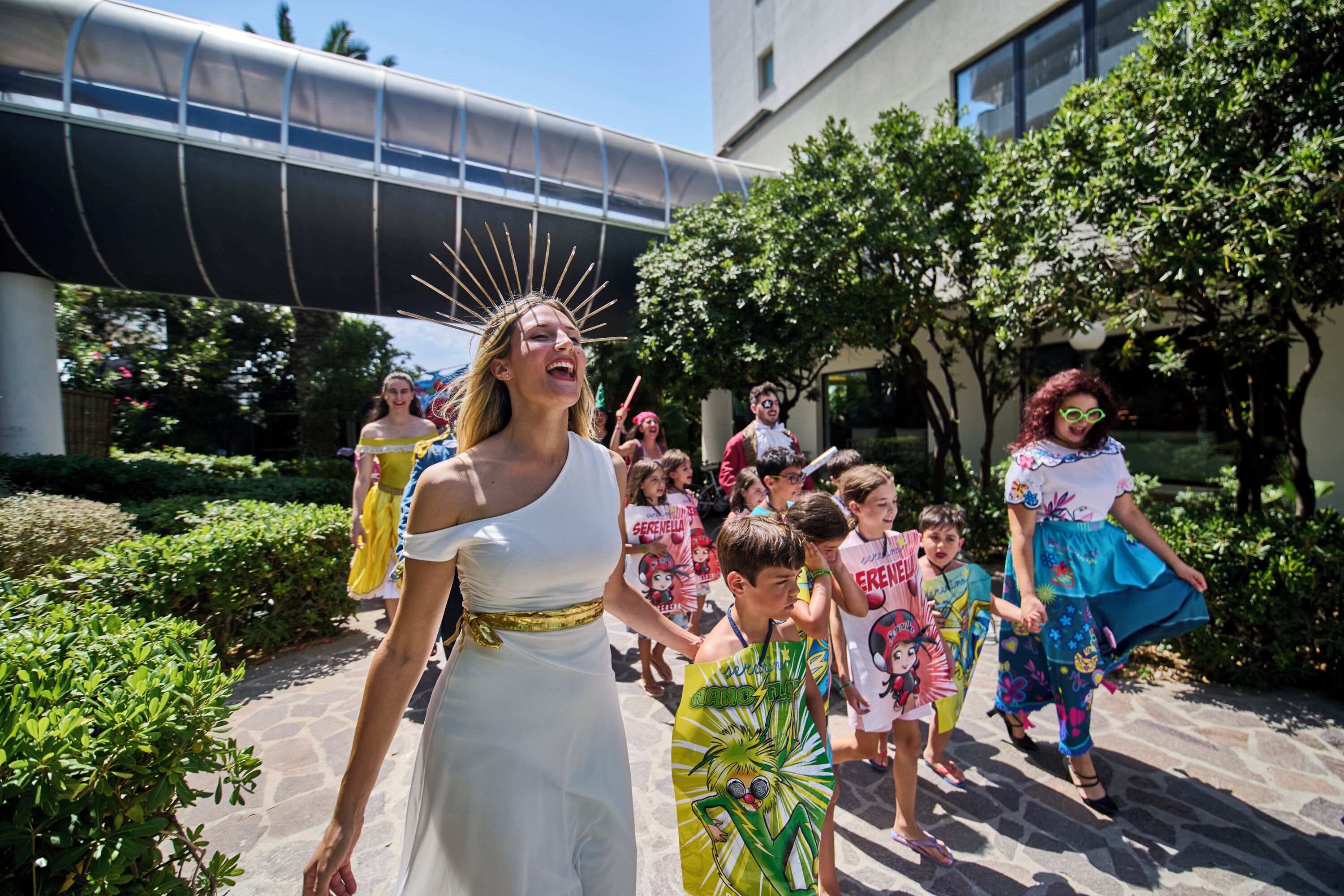 A group of people walking outdoors, some in colorful costumes, with greenery and a building in the background.