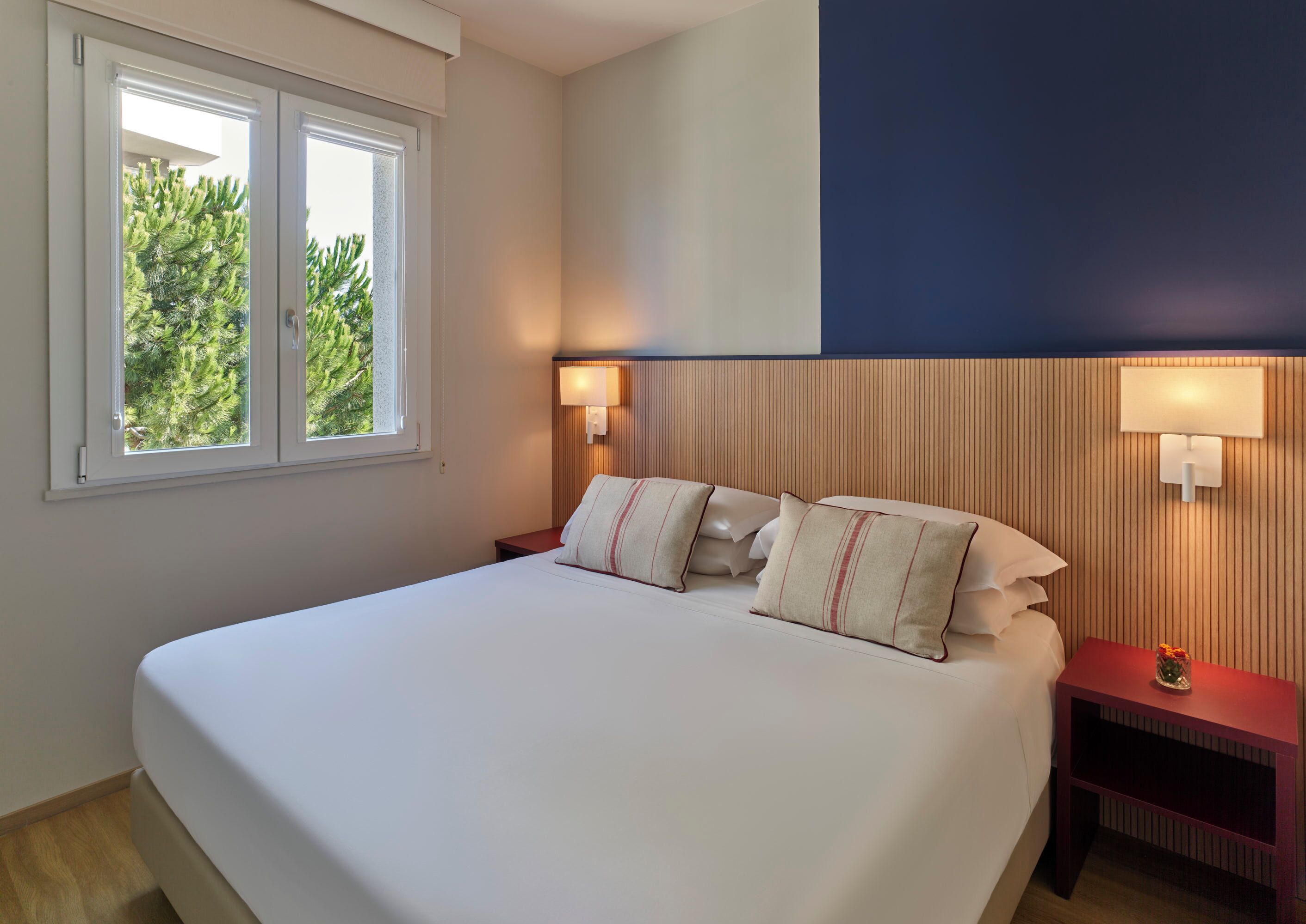 A hotel room with a large bed, two bedside tables with lamps, a window with a view of trees, and a wall with vertical wooden paneling.