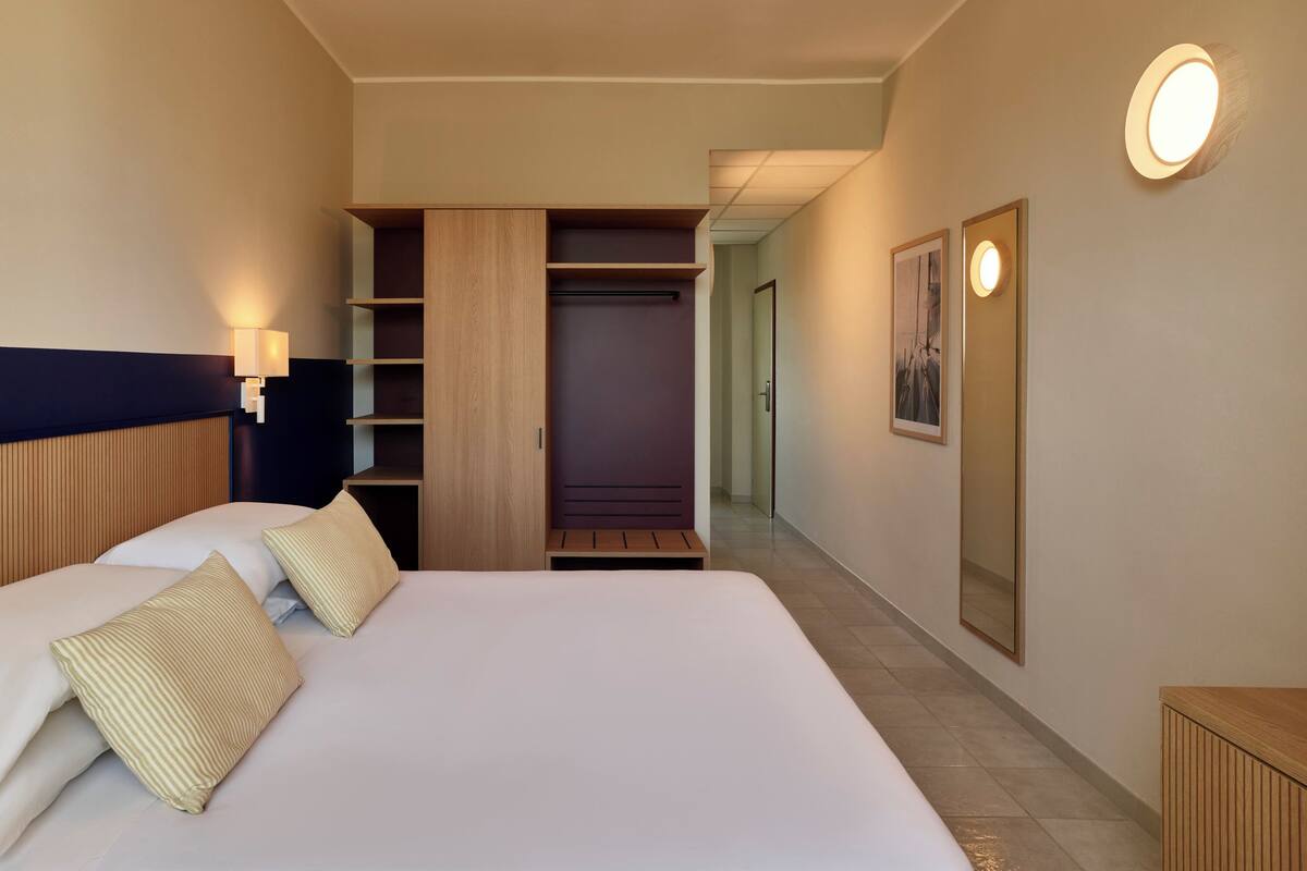 A hotel room with a large bed, a wardrobe, a mirror, and a hallway leading to another room.