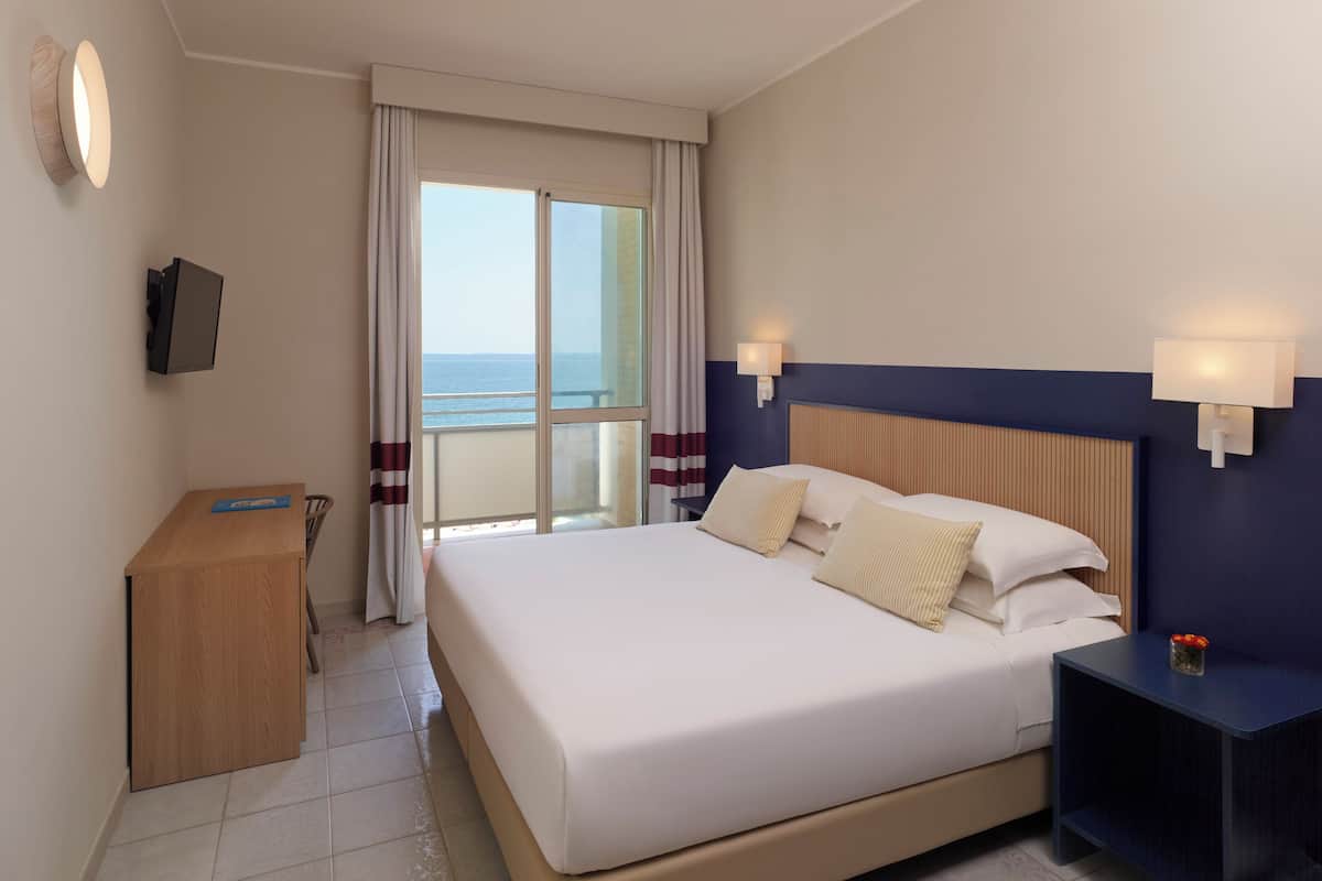 A hotel room with a bed, a desk, a TV, and a view of the sea.