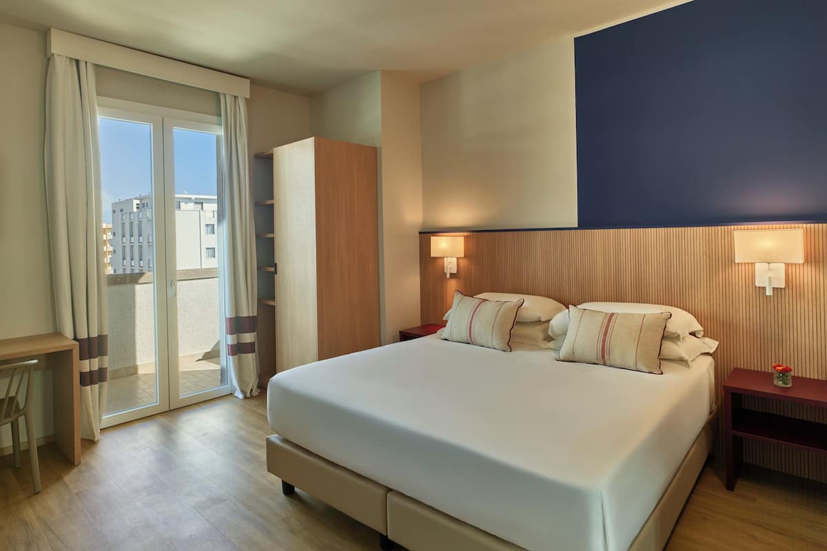 A hotel room with a bed, a desk, a chair, a nightstand, a lamp, and a view through a sliding door.