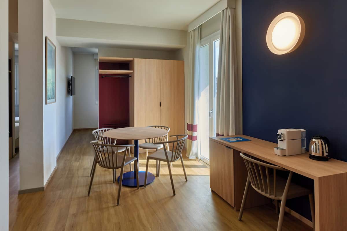 A modern room with a wooden dining table and chairs, a small kitchenette, and a large window with curtains.