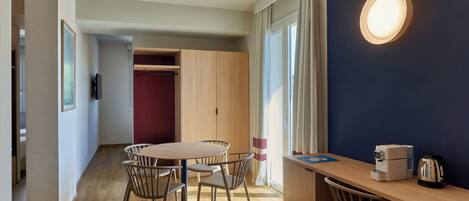 A modern room with a wooden dining table and chairs, a small kitchenette, and a large window with curtains.