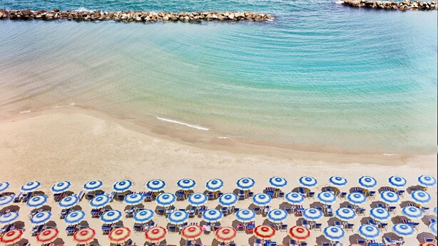 A beach with rows of blue and red umbrellas and chairs, a rocky breakwater, and calm turquoise water. - Serena Majestic Hotel (Montesilvano)