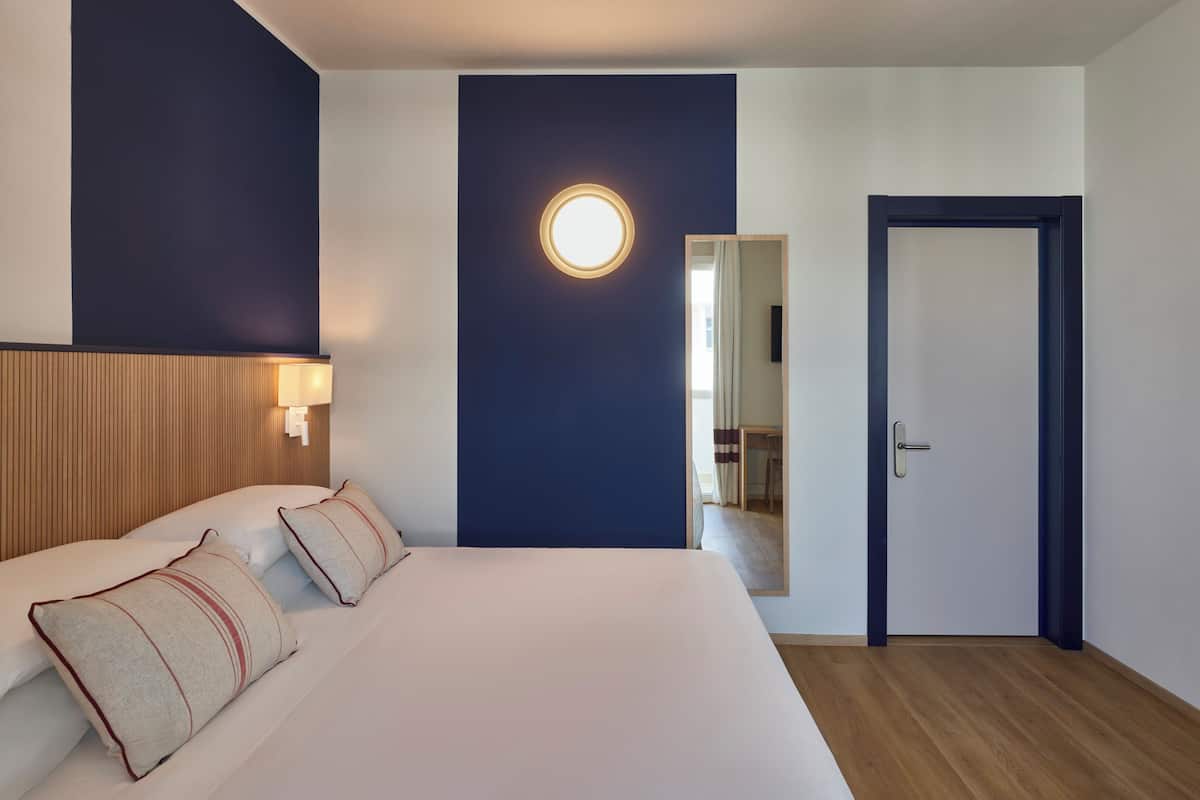 A hotel room with a large bed, a wooden headboard, a round wall light, a mirror, and a door.