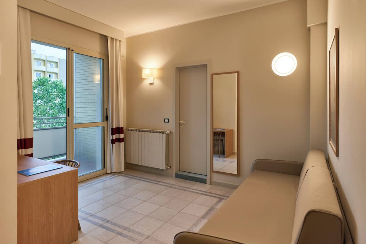 A hotel room with a large window, a radiator, a desk, a chair, and a sofa.