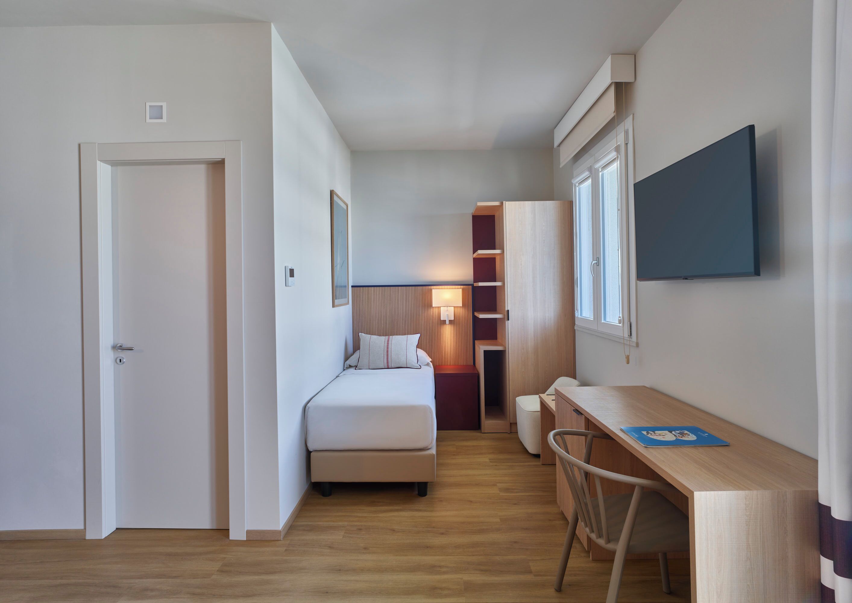 Una habitación de hotel pequeña con cama, mesita de noche, escritorio, silla y televisor montado en la pared.