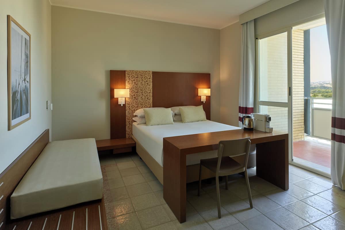 A hotel room with a bed, bedside table, chair, and a view of the outdoors through a large window.