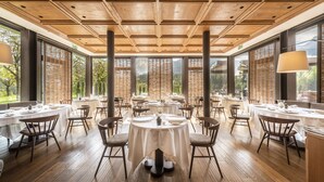 Un vaste espace dédié à la salle à manger, avec des tables et des chaises rondes, de grandes fenêtres donnant sur un paysage verdoyant et un plafond en bois.
