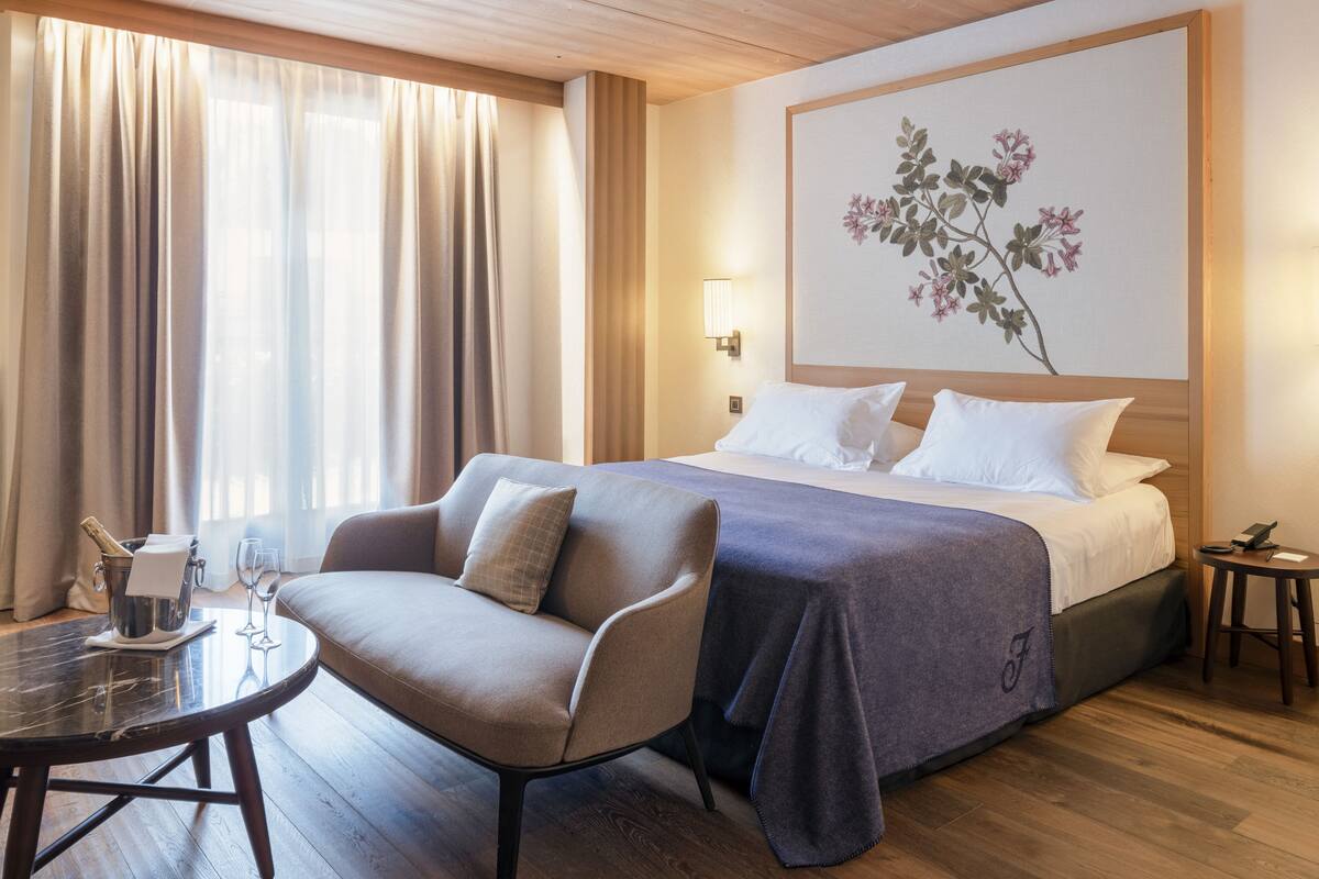 A hotel room with a bed, a sofa, a small table with a bottle and glasses, and a painting on the wall.
