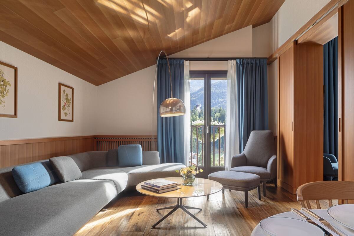 A modern living room with a wooden ceiling, a sofa, a chair, a round coffee table, and a view of mountains through a glass door.