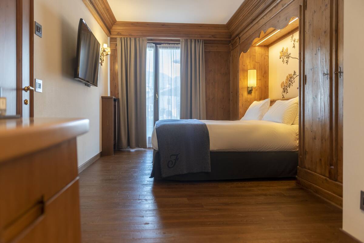 A hotel room with a wooden floor, a bed with a gray blanket, a TV mounted on the wall, and a window with curtains.