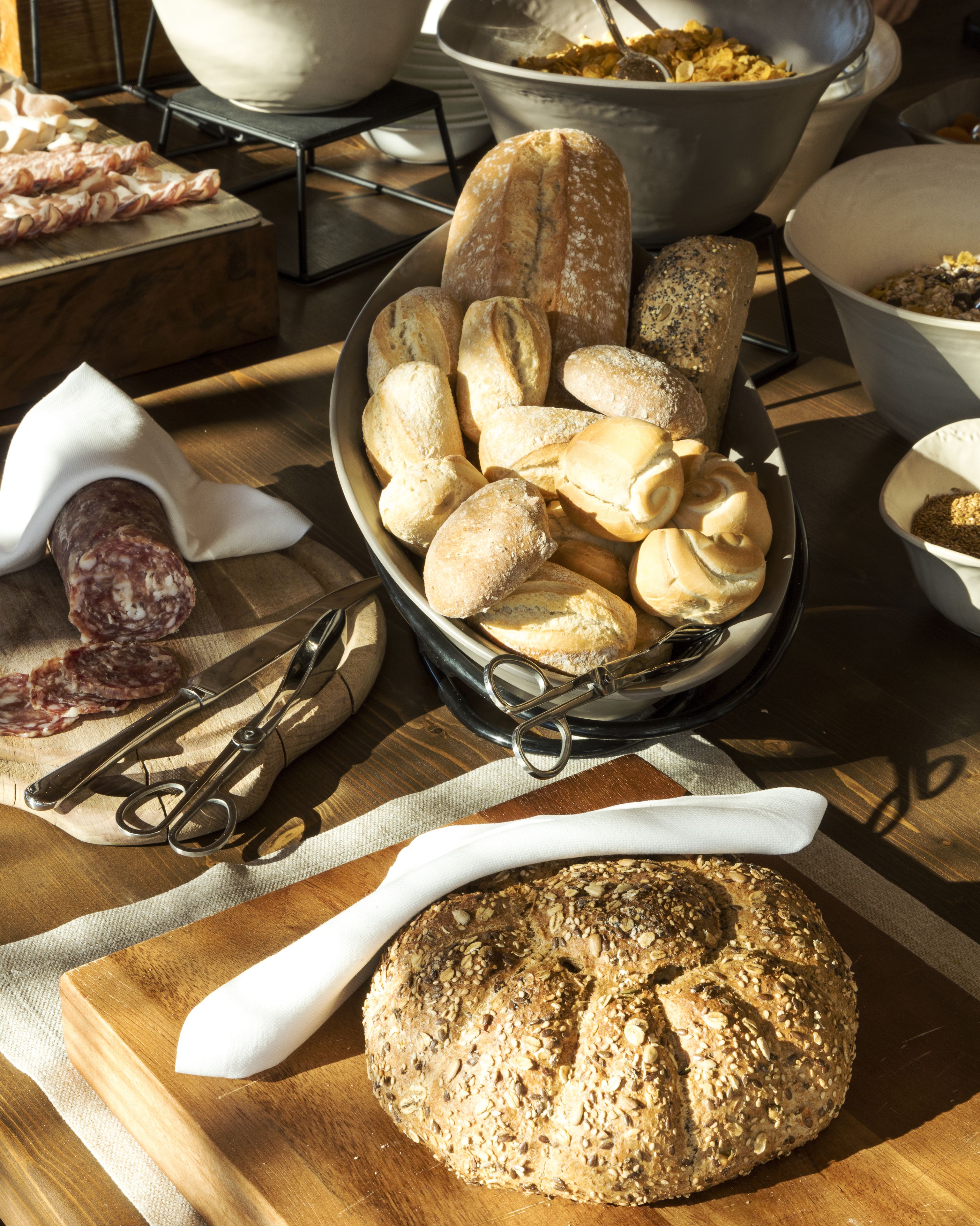 Verschiedene Brotsorten und Gebäck auf einem Holztisch, im Vordergrund ein Roggenbrot und eine Schüssel mit Brotscheiben.