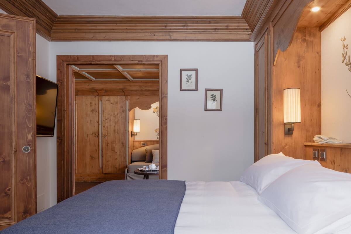 A hotel room with a wooden door, a bed with white linens, a small table with a cup, and a wall-mounted lamp.