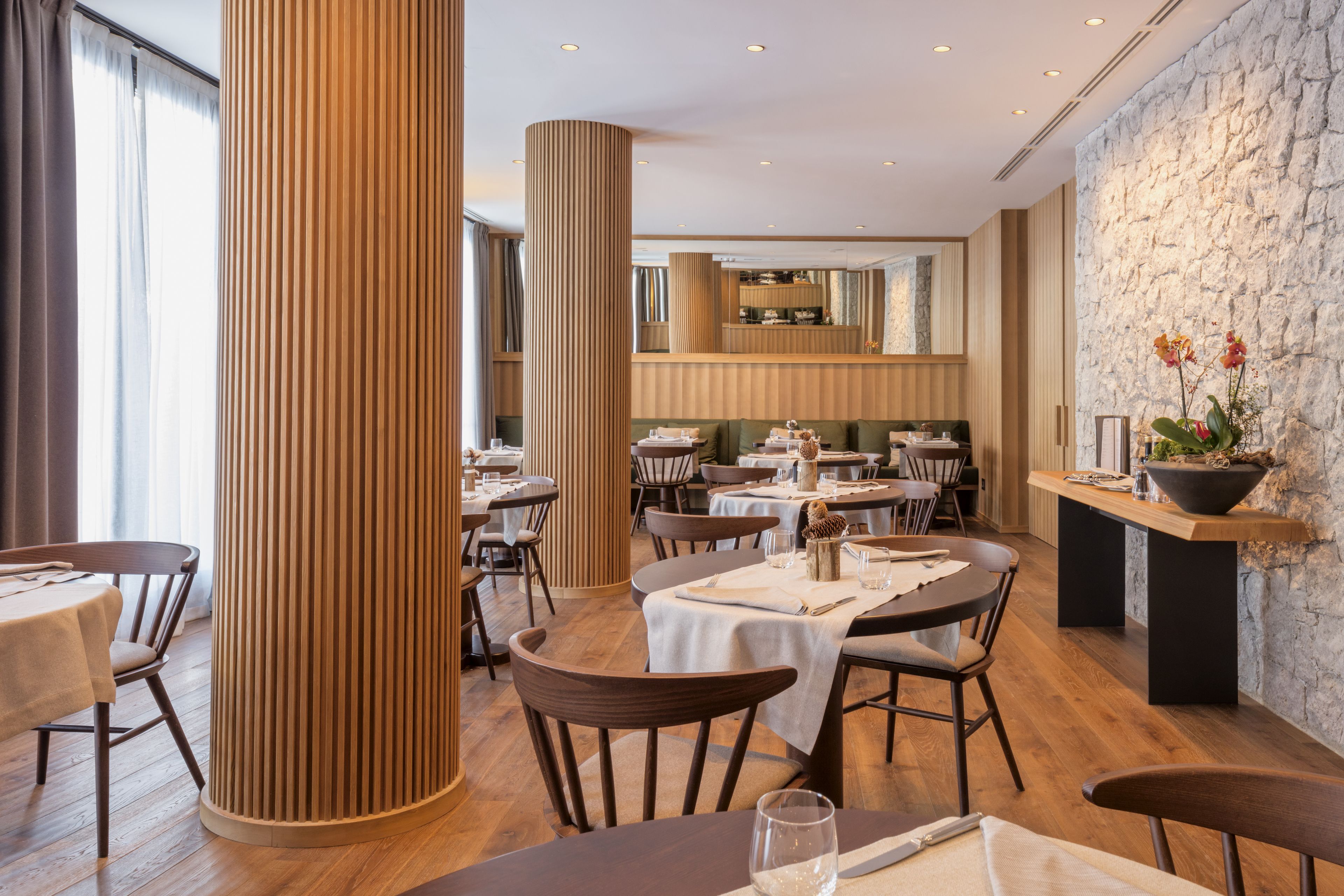 A restaurant with wooden floors, round tables, and chairs. There are large cylindrical columns and a stone wall.