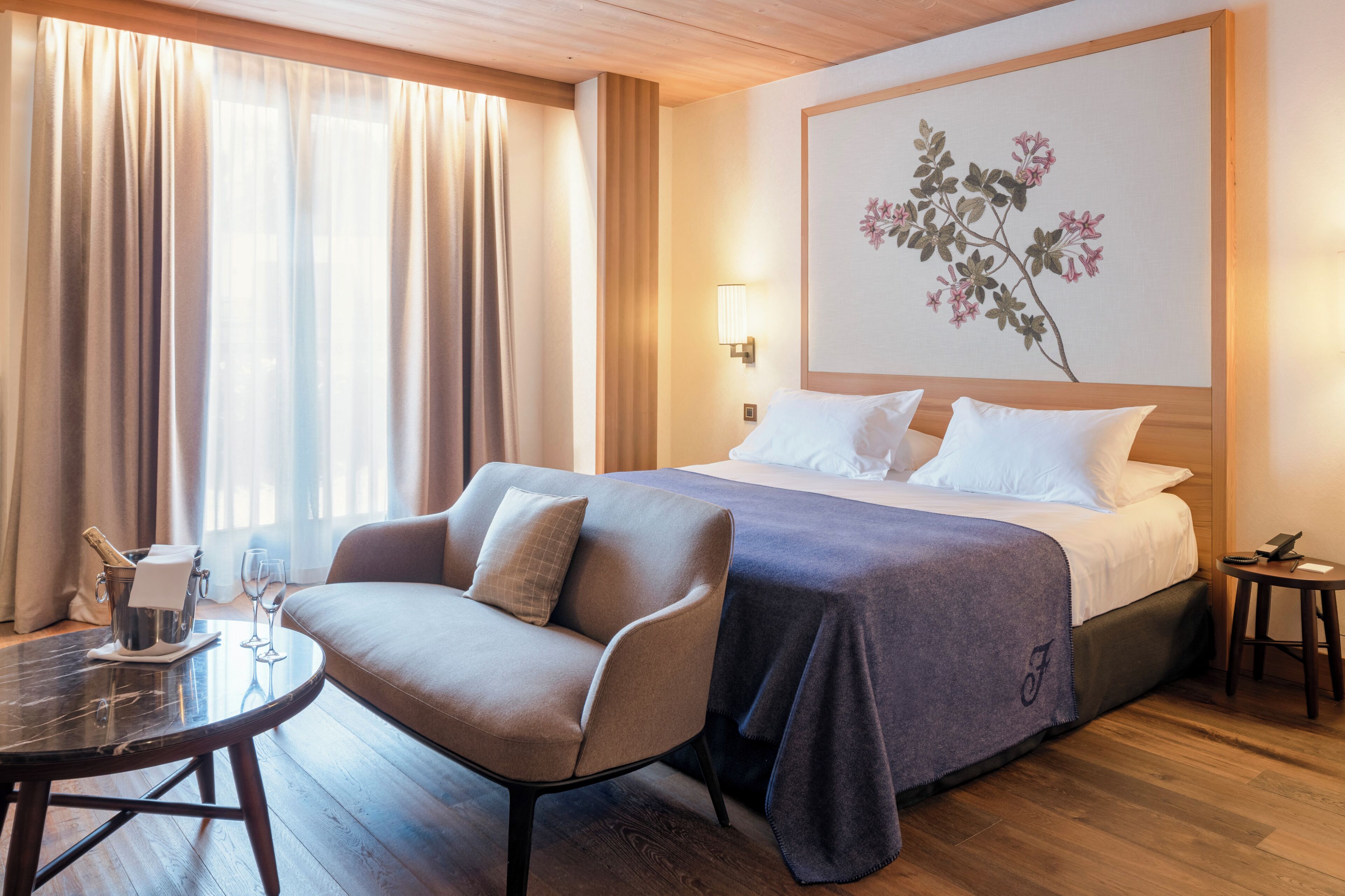 A hotel room with a bed, a sofa, a small table with a bottle and glasses, and a painting on the wall.