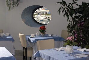 A dining area with round tables, chairs, and a view of boats through a circular window. - Hotel Villa Carolina (Forio)
