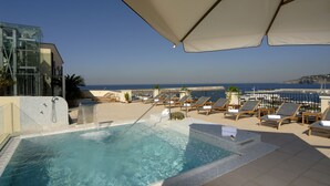 Una piscina en la azotea con agua cristalina, rodeada de sillones de descanso y sombrillas, con vistas a un puerto deportivo y al mar.