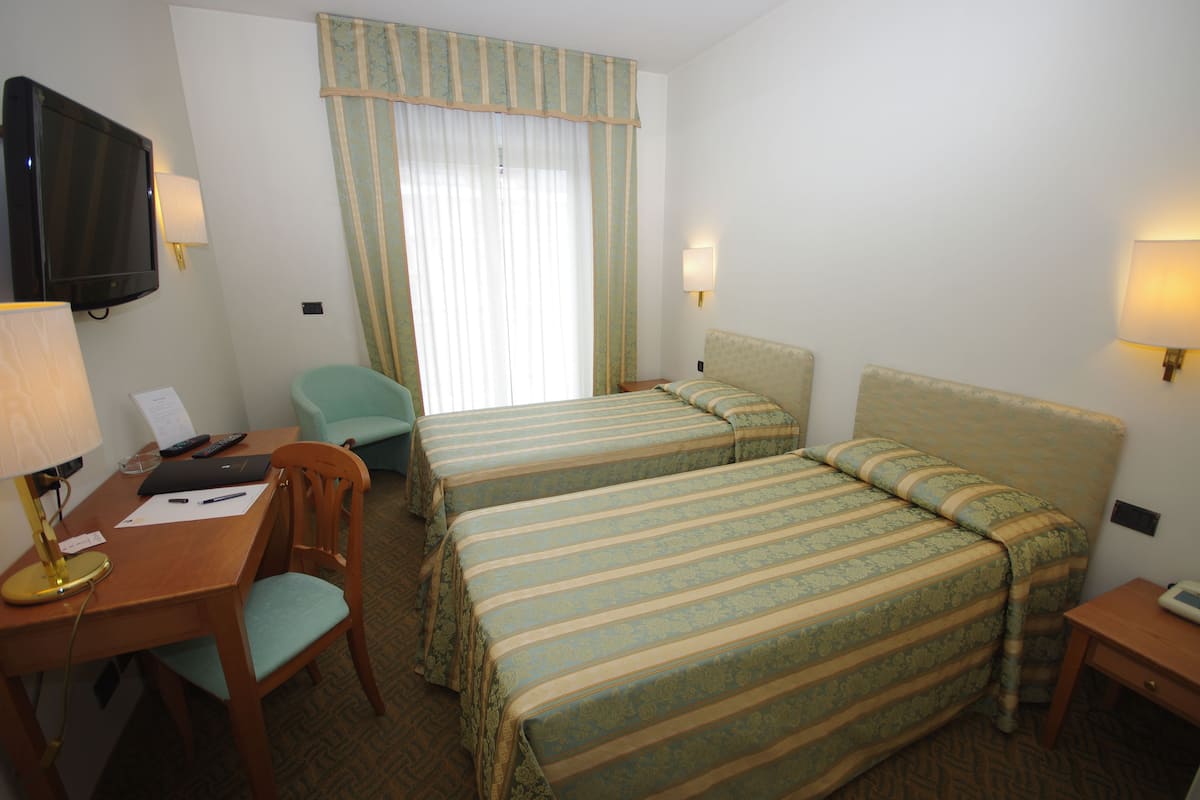 A hotel room with two beds, a desk with a lamp, a chair, a television, and a window with curtains.