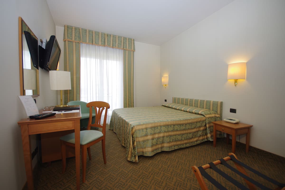 A hotel room with a bed, a desk with a chair, a lamp, and a bedside table.