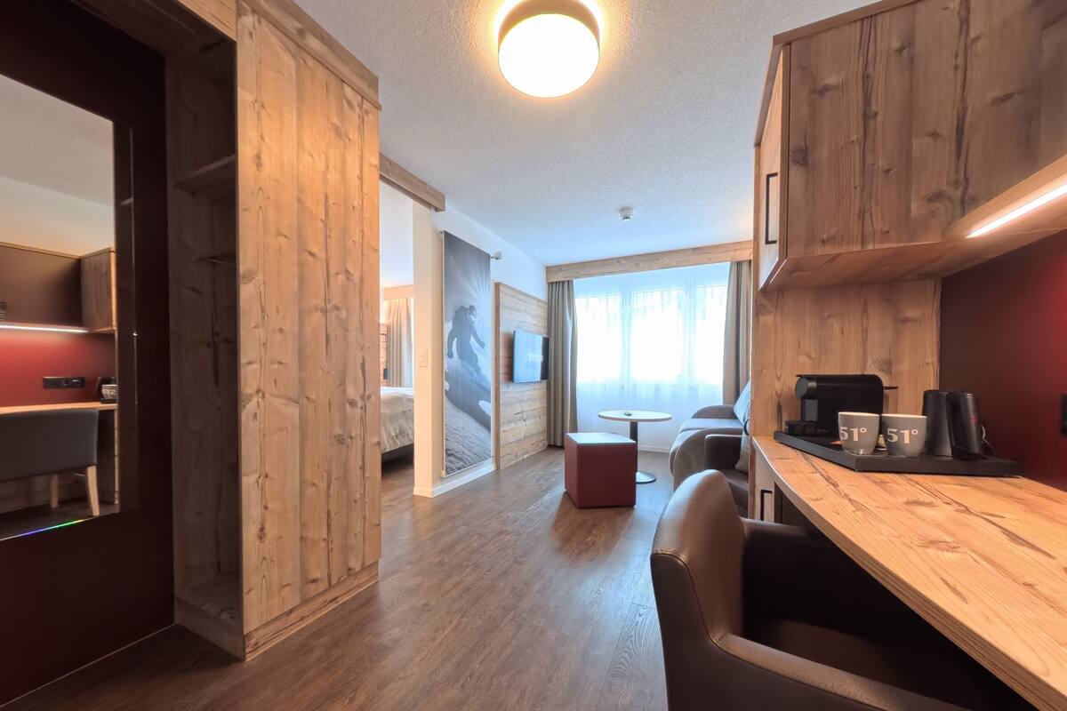 A modern hotel room with wooden accents, a cozy seating area, a small table, and a large window with curtains.