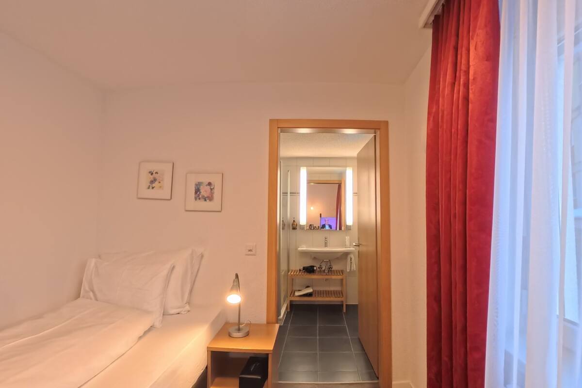 A hotel room with a bed, a nightstand with a lamp, a bathroom with a mirror, and a red curtain.