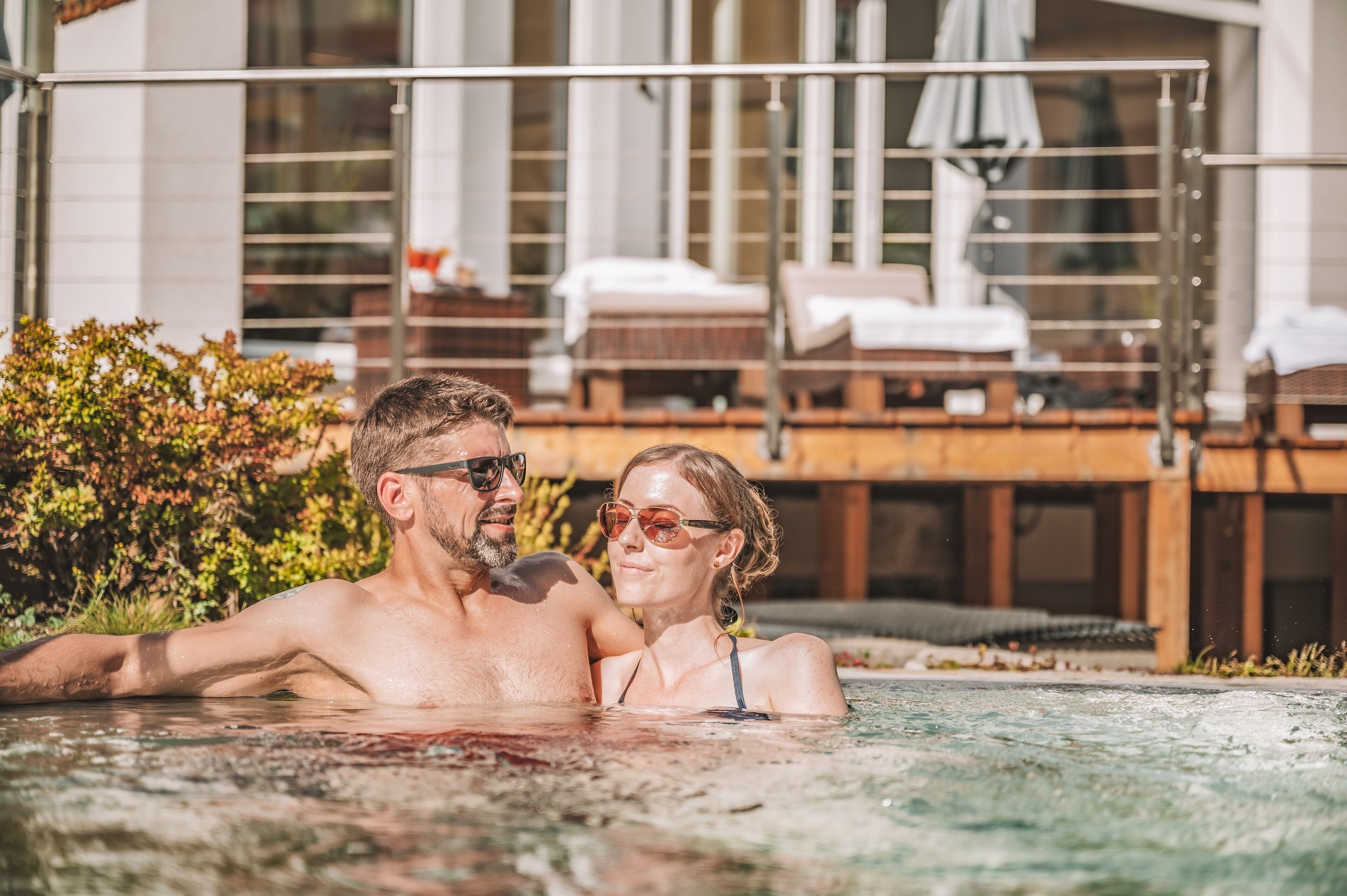Una pareja en un jacuzzi con una terraza de madera y sillones de descanso al fondo.