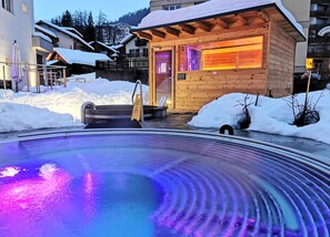 Une cabane en bois avec une porte en verre, entourée de neige, et une piscine éclairée de lumières violettes.