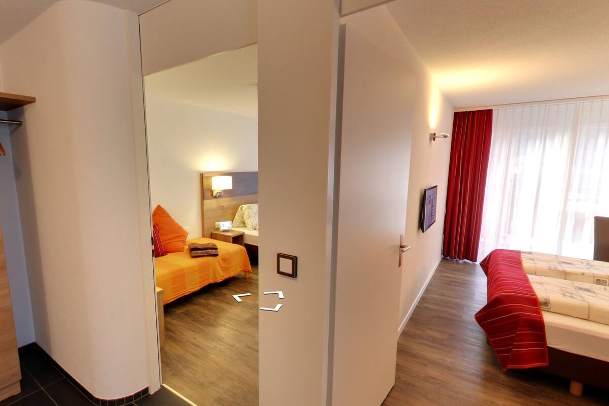 A hotel room with a wooden wardrobe, a bed with a red blanket, a bedside table with a lamp, and a large window with curtains.