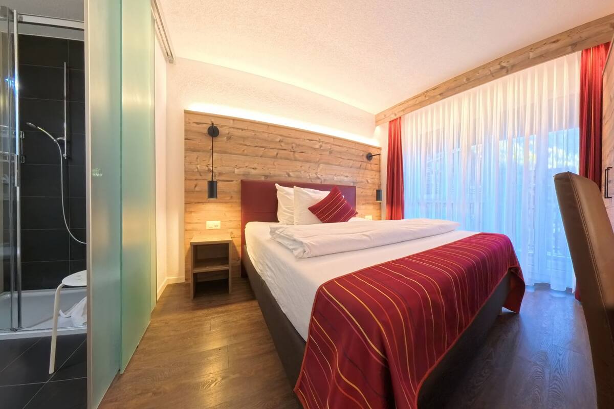 A hotel room with a wooden headboard, a bed with a red-striped bedspread, a nightstand, a wooden desk, and a shower area.