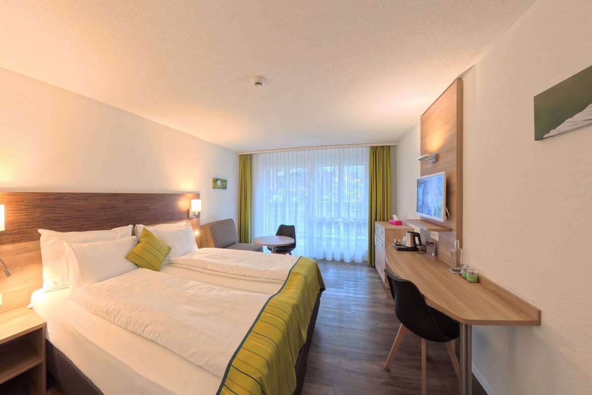 A hotel room with a bed, bedside table, lamp, desk, chair, and a TV.