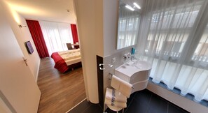 Une chambre d’hôtel moderne avec un plancher en bois, un lit avec des rideaux rouges, un lavabo blanc et un grand miroir.