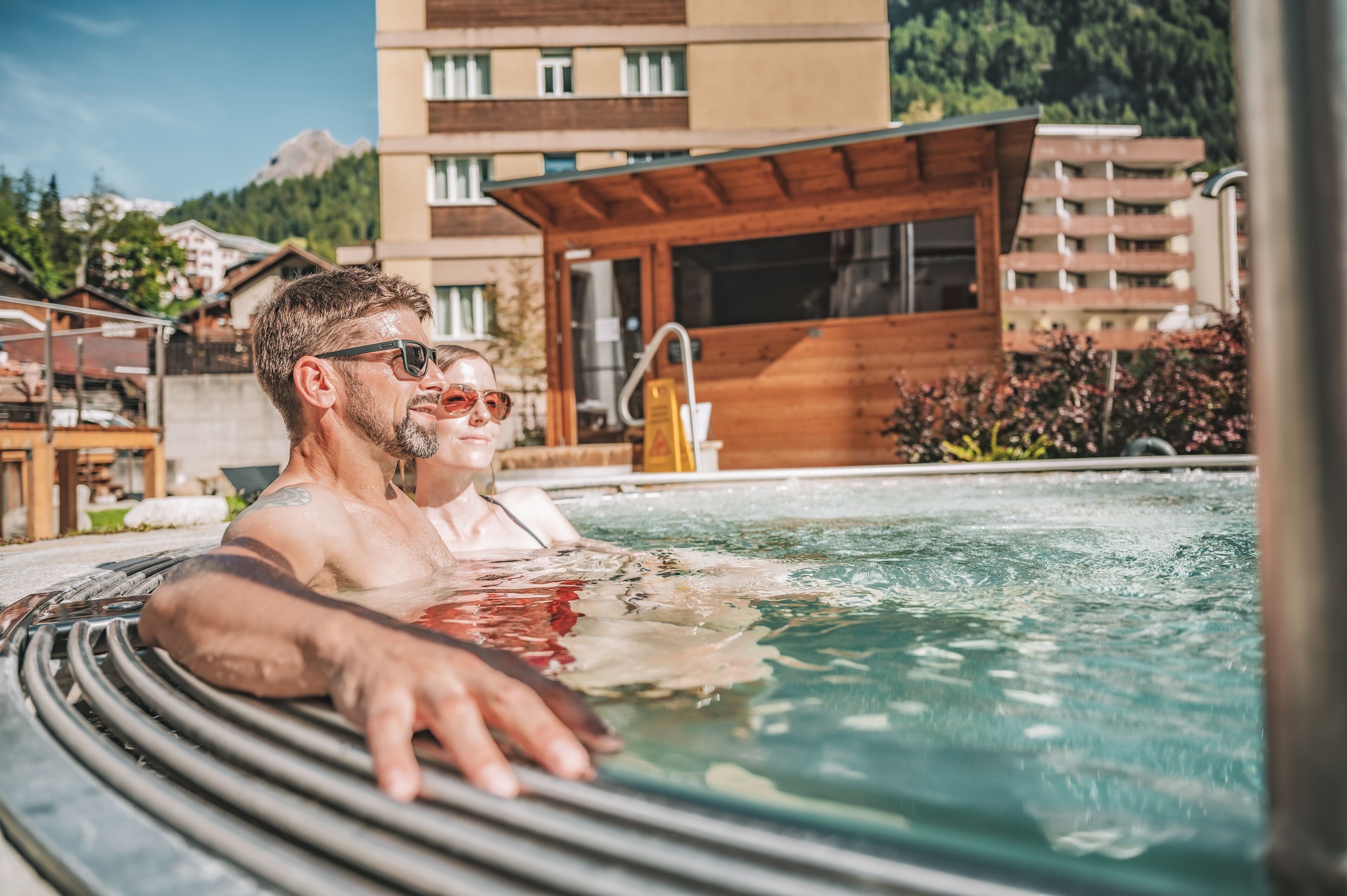 A man in a hot tub with a wooden deck, surrounded by buildings and greenery.