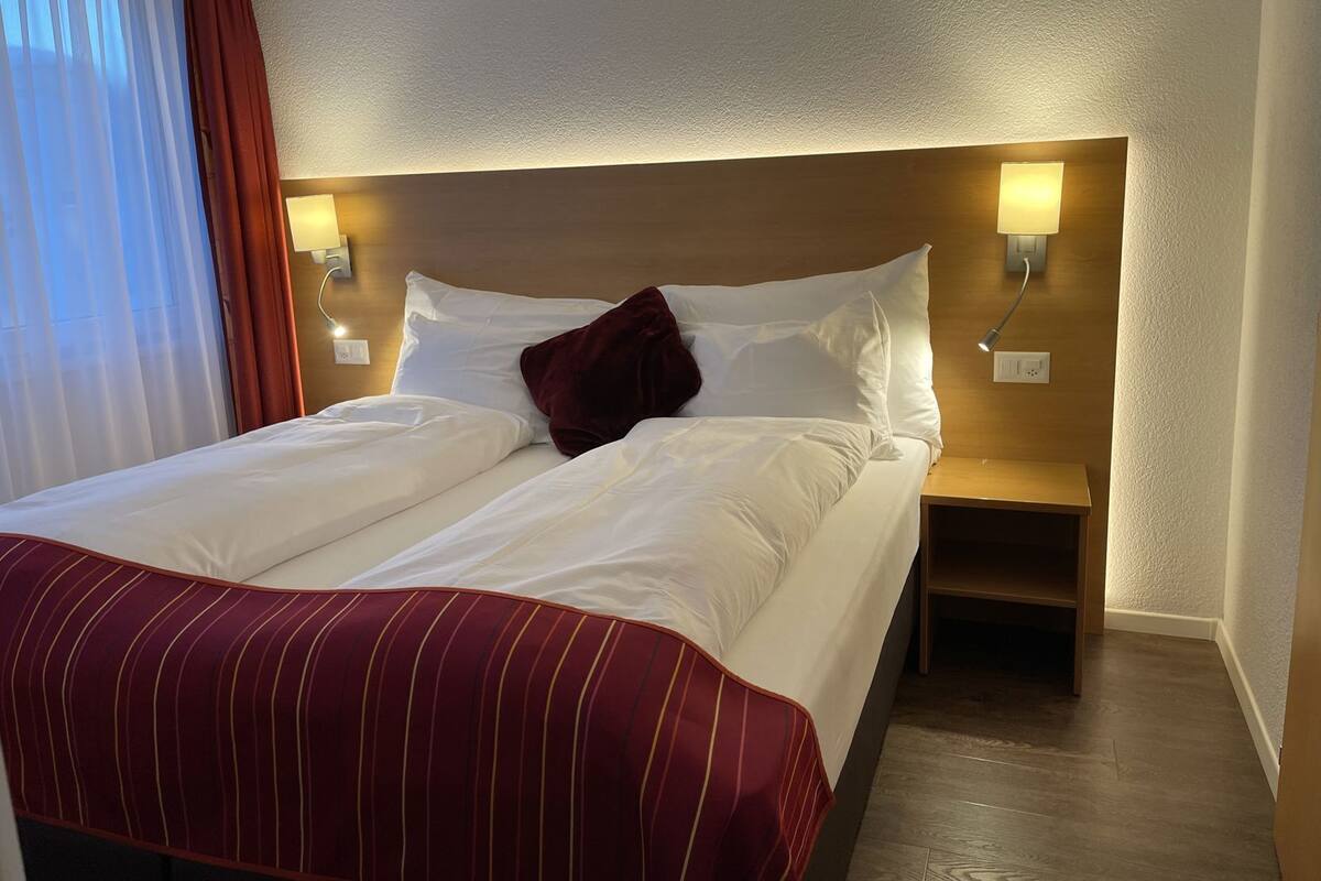 A hotel room with a double bed, two bedside tables, a headboard with integrated lighting, and a window with curtains.