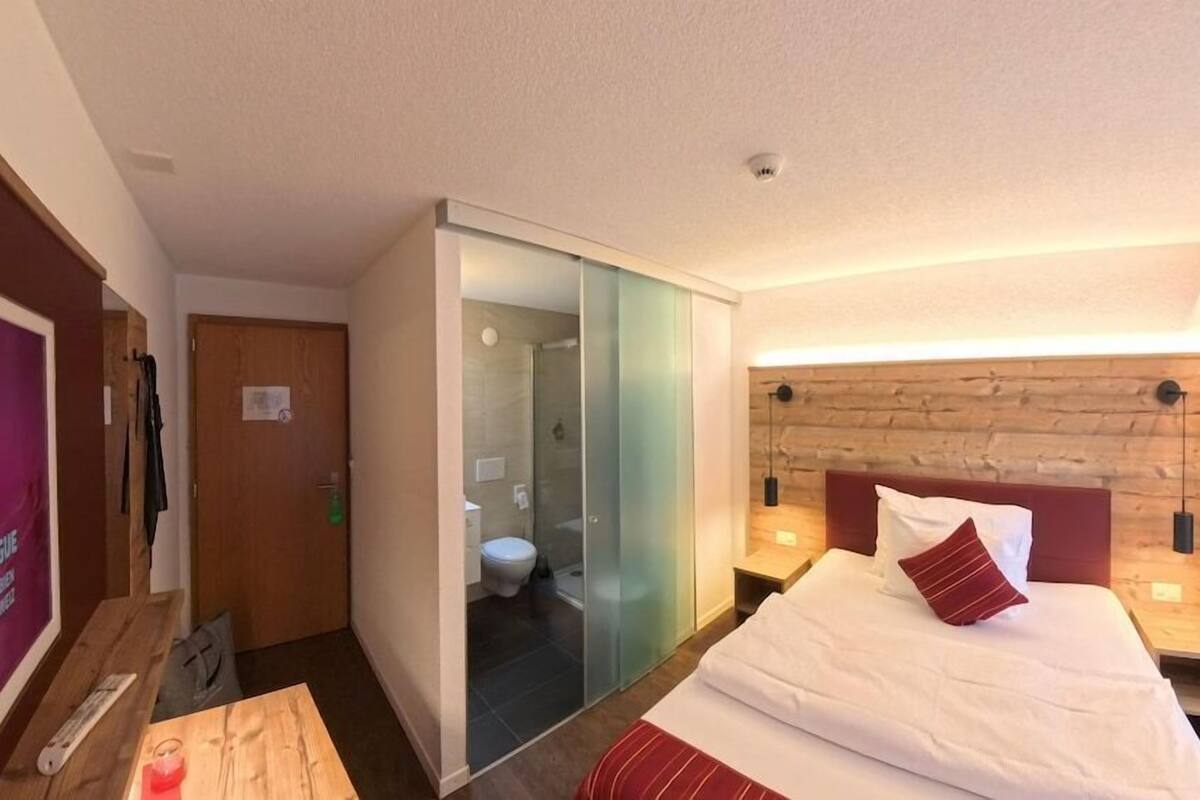 A hotel room with a bed, a wooden headboard, a nightstand, a desk, and a glass partition separating the room from the bathroom.