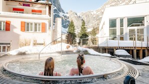 A hot spring area with two people relaxing in the water, surrounded by snow-covered mountains and buildings with red shutters.
