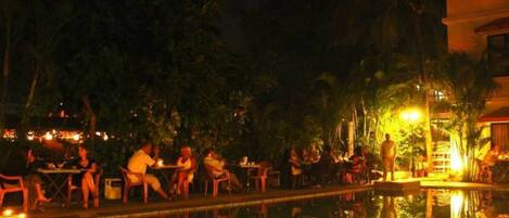 An outdoor pool area with tables and chairs, surrounded by trees and a building in the background.