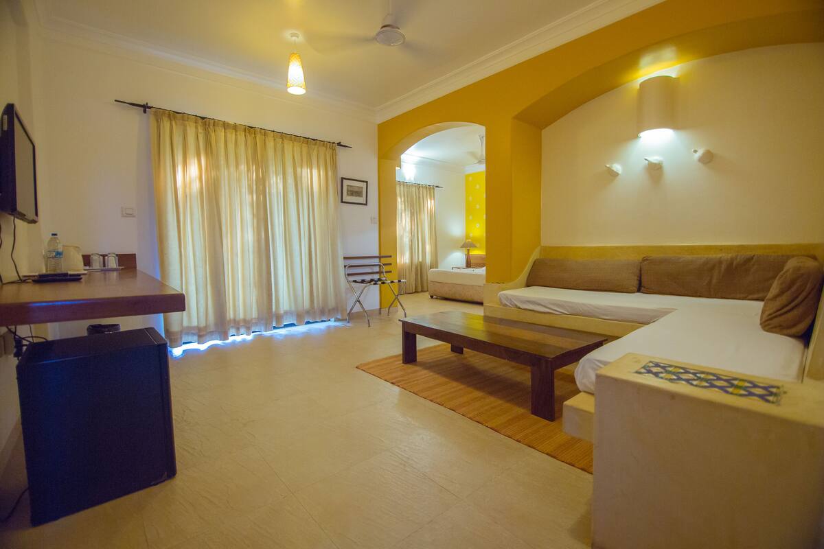 Habitación de hotel con cama, escritorio, silla y una mesita. Se ve una ventana con cortinas y vista a otra habitación.