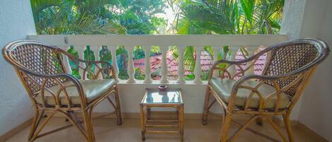 A balcony with two wicker chairs and a small table, overlooking a lush garden with palm trees.