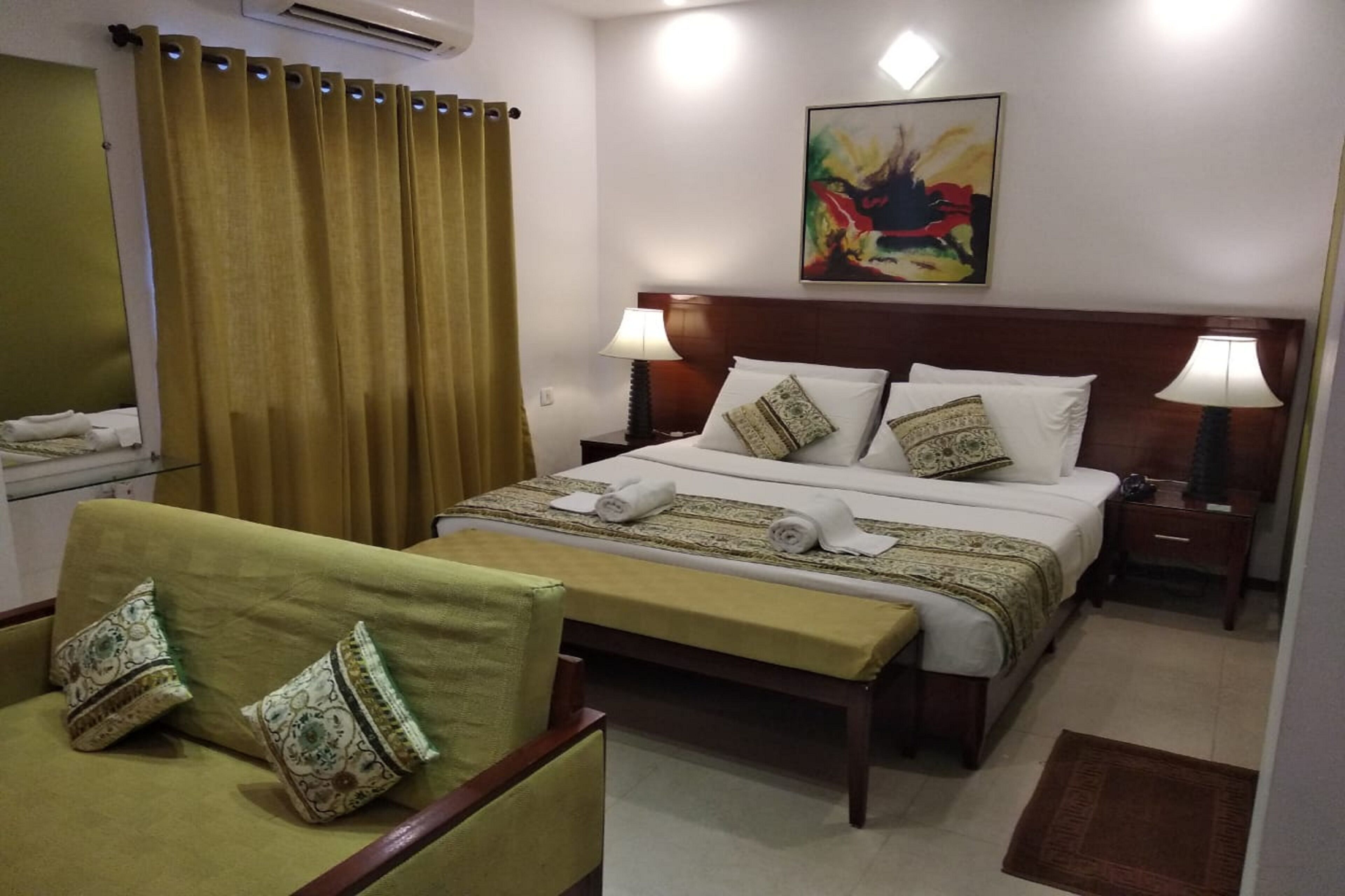 Habitación de hotel con cama, dos mesitas de noche, una silla, un banco, un ventanal con cortinas y un cuadro en la pared.