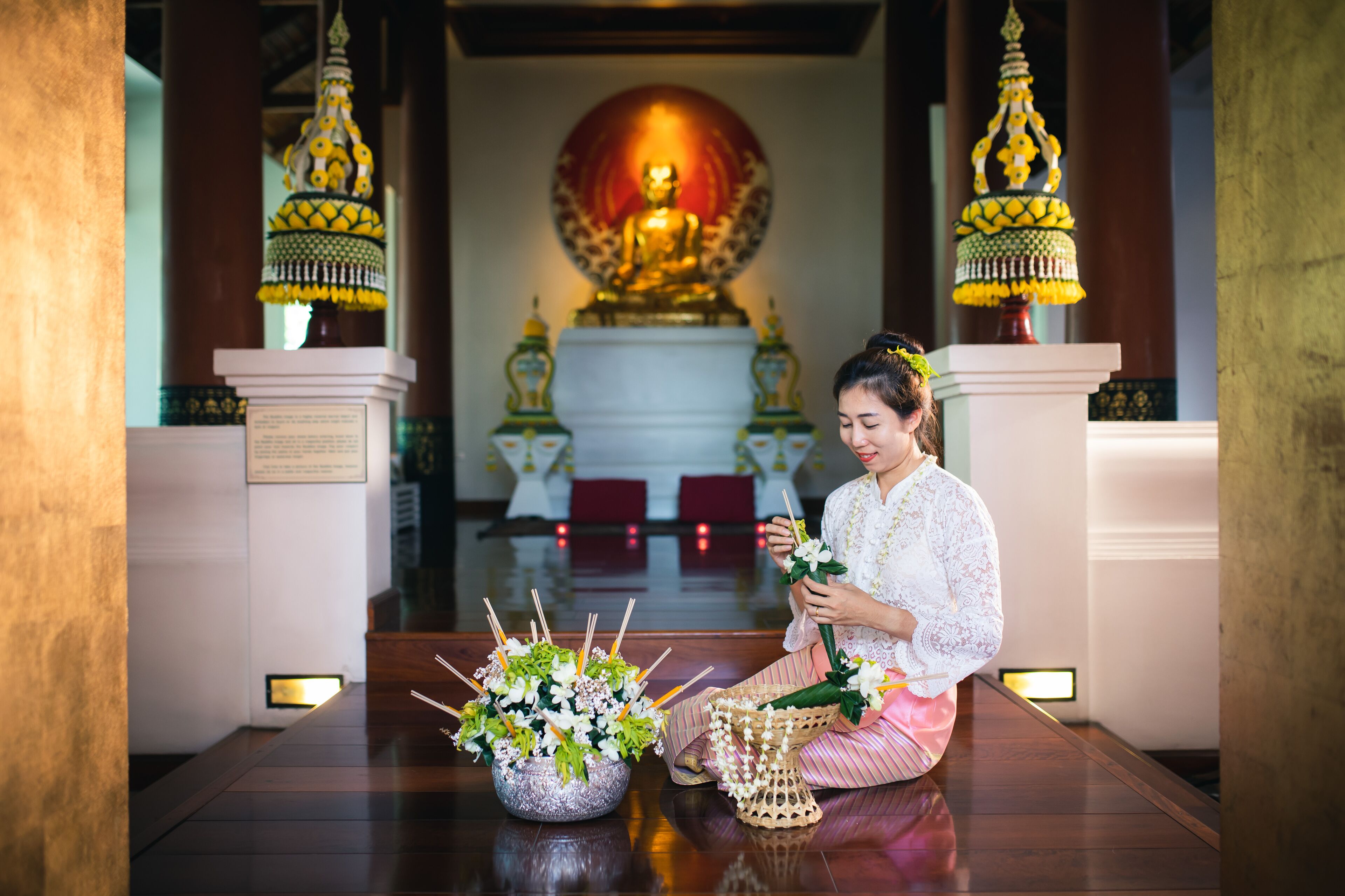 A person sitting on the floor in a room with a Buddha statue in the background, a flower arrangement, and traditional decorations.