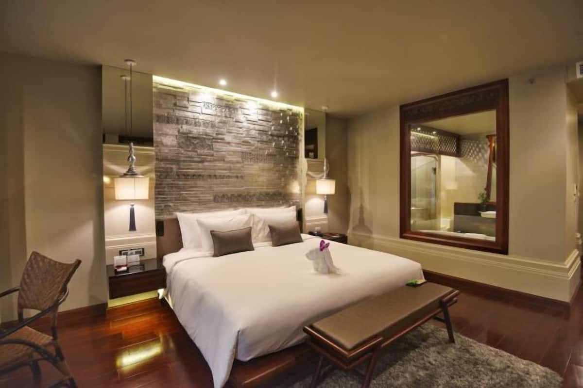 Quarto de hotel com uma cama grande, luminárias laterais, cabeceira de madeira, um banquinho e um espelho.