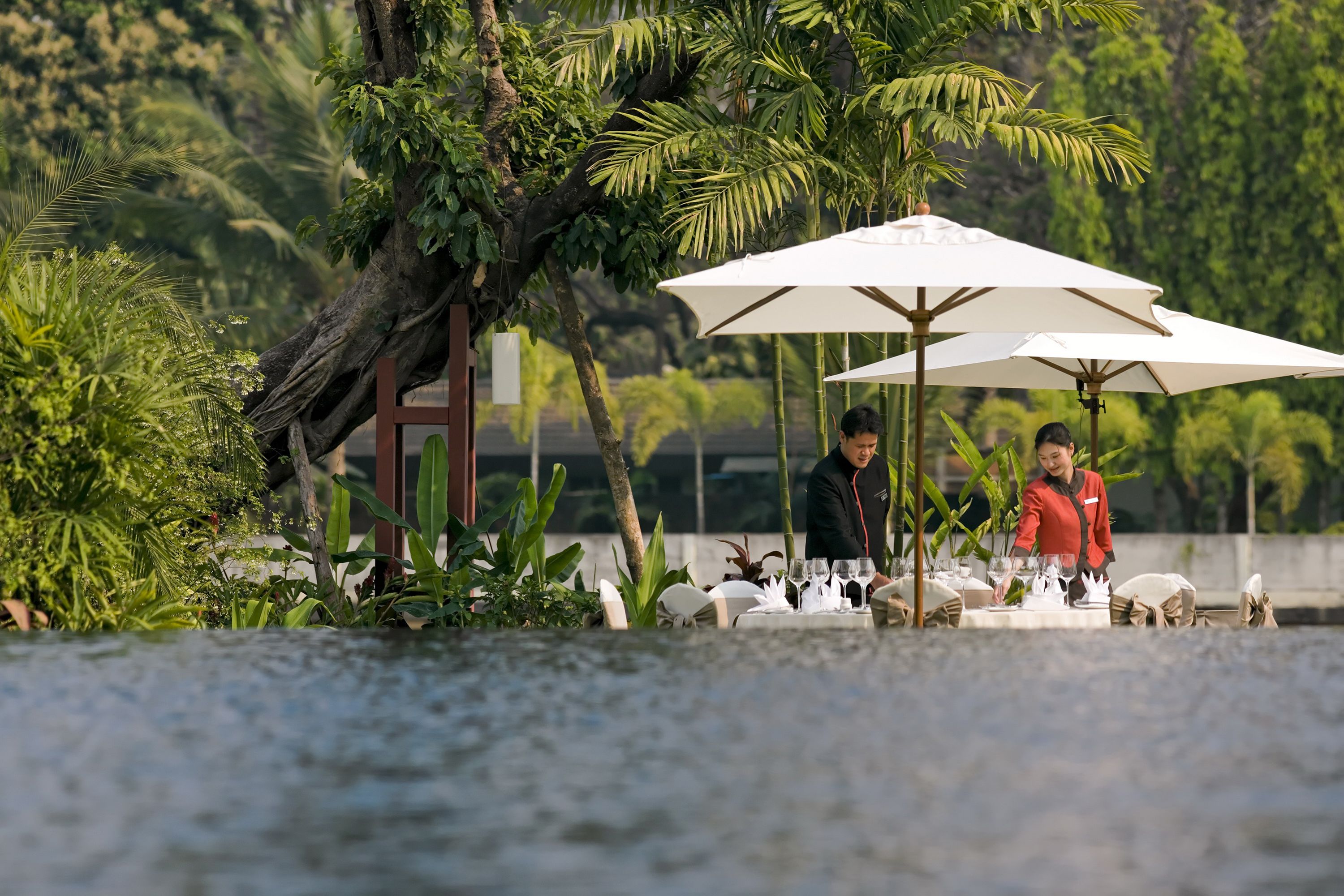 An outdoor dining area with tables set for two, surrounded by lush greenery and a large umbrella providing shade.