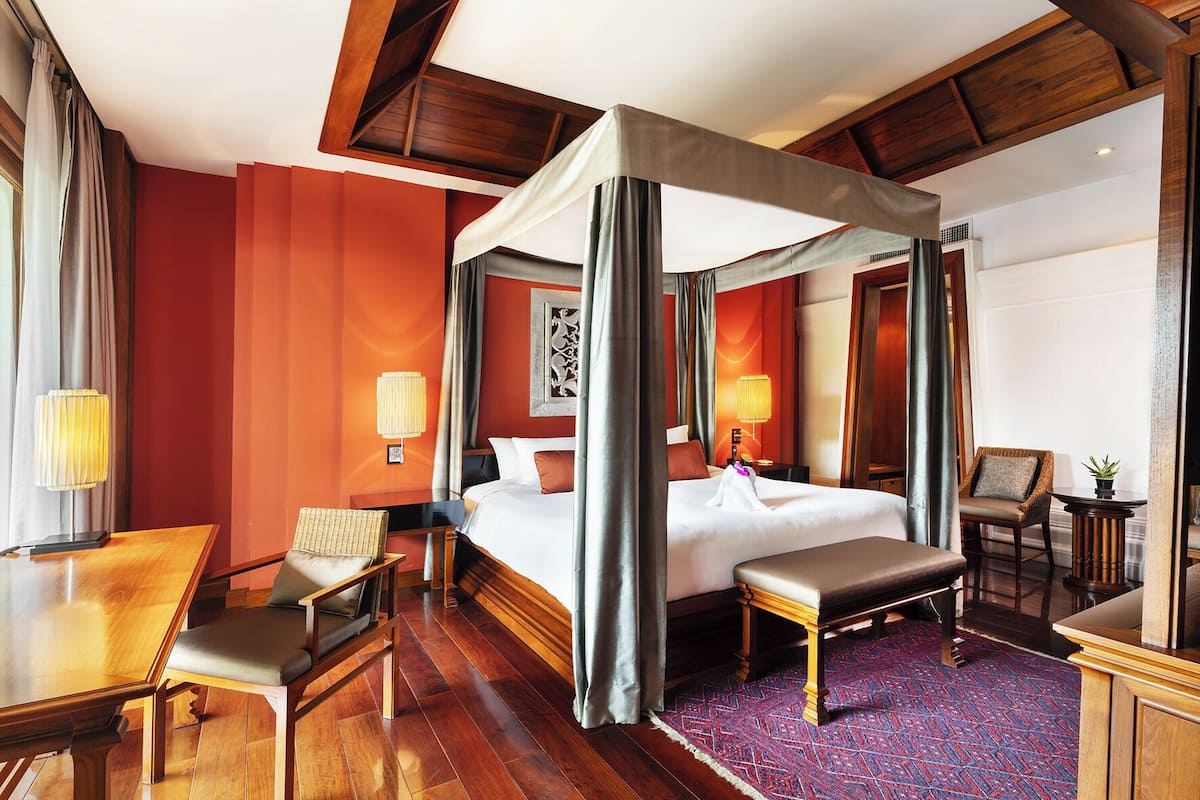 Quarto de hotel com cama de dossel, uma escrivaninha de madeira, uma cadeira, um banco e uma pequena mesa com uma planta.